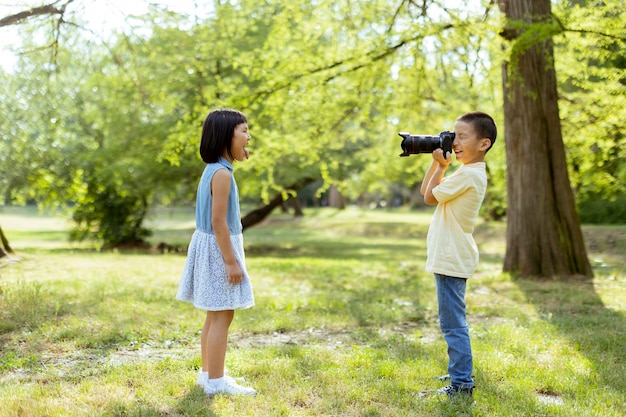 Kleiner asiatischer Junge, der sich wie ein professioneller Fotograf verhält, während er Fotos von seiner kleinen Schwester macht