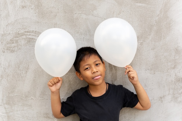 Kleiner asiatischer Junge, der weiße Ballone über grauem konkretem Hintergrund hält.