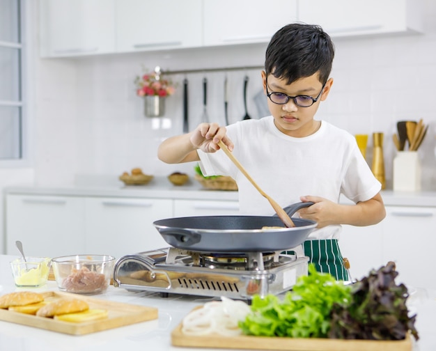 Kleiner asiatischer Junge in Gläsern, der Pasteten auf heißer Pfanne braten, während er Burger zum Mittagessen in der modernen hellen Küche zu Hause zubereitet. Konzept für Bildung und Lernen durch Handeln.