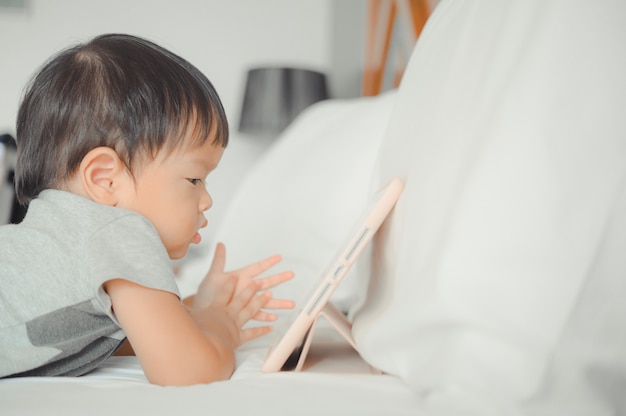 Kleiner asiatischer Junge liegt im Bett und sieht sich eine Geschichte auf dem Touchscreen-Tablet an