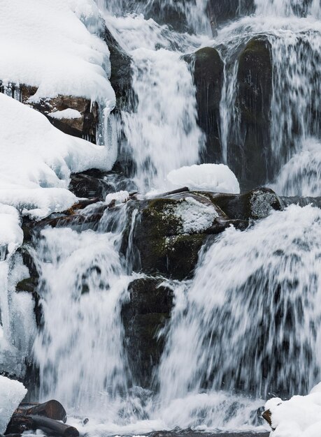 Kleiner Bergwasserfall aus eisigem Wasser fließt zwischen nassen Steinen, die mit weißem Schnee bedeckt sind