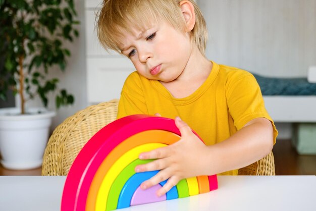 Kleiner blonder Haarjunge in Kleidung aus natürlichem Stoff spielt regenbogenfarbenes Holzspielzeug am weißen Tisch Eco