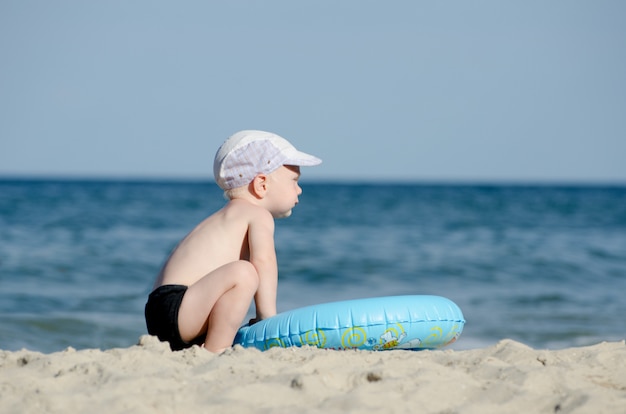 Kleiner blonder Junge, der an der Küste mit einem Schwimmkreis sitzt