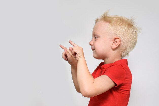 Kleiner blonder Junge im roten T-Shirt zeigt mit dem Finger. Platz für Text. Platz für Werbung. weißer Hintergrund