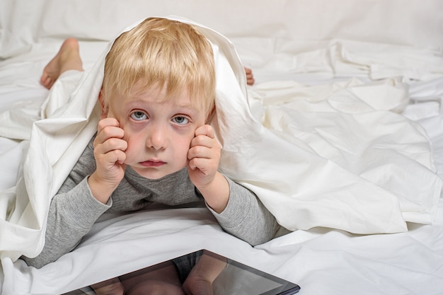 Kleiner blonder Junge mit müden Augen von der Tablette. Im Bett liegen und sich unter der Decke verstecken. Gadget Freizeit