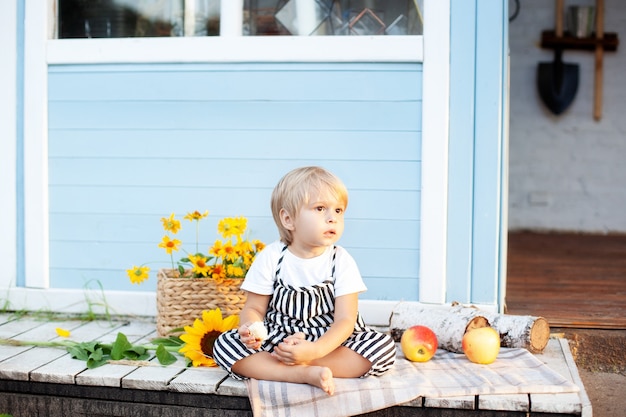 Kleiner blonder Junge sitzt zu Hause auf einer Holzveranda Glückliches Kind spielt im Sommerhof