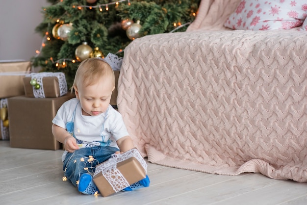 Kleiner blonder Junge spielt mit einer hellen Girlande am Weihnachtsbaum in gemütlichem Zuhause.