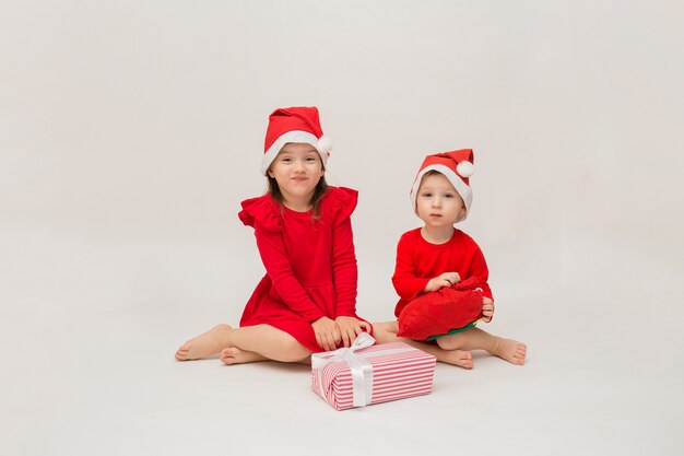 Kleiner Bruder und Schwester in Weihnachtskostümen mit Mützen mit Geschenken an einer weißen Wand