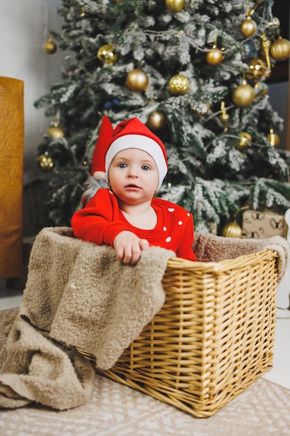 kleiner einjähriger Junge neben dem Weihnachtsbaum eine Idee für ein Kinderfoto für das Neujahrsstudio Neujahrsdekoration Weihnachtsgeschenke rotes festliches Kinderkostüm