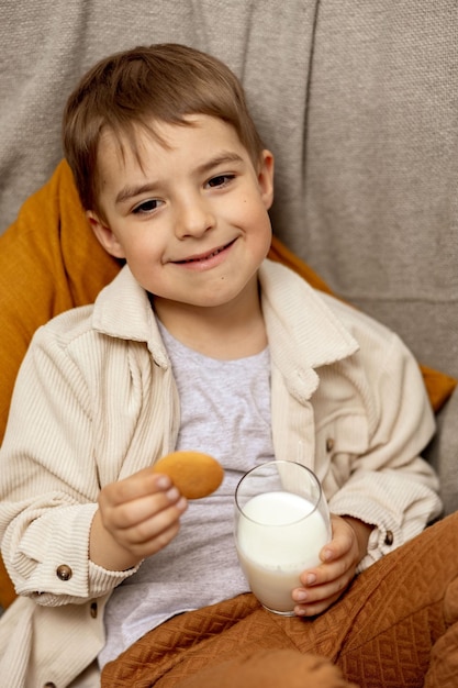 Kleiner entzückender Junge, der zu Hause auf der Couch sitzt und Milch mit Cookie trinkt Frische Milch im Glas Molkerei gesundes Getränk Healthcare-Quelle von Calcium-Laktose Vorschulkind mit Freizeitkleidung