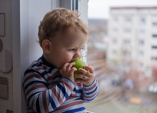 Kleiner europäischer Junge isst einen grünen Apfel, der auf der Fensterbank im Raum sitzt