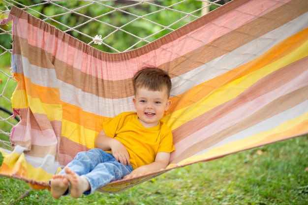 Kleiner glücklicher Junge, der in einer hellen Hängematte in seinem Park reitet. Sommer Kinderferien