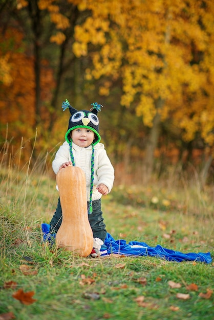 Kleiner glücklicher Junge in einer gestrickten Mütze und in warmen Kleidern, die mit Kürbis auf Rasen zwischen den Herbstbäumen bleiben.