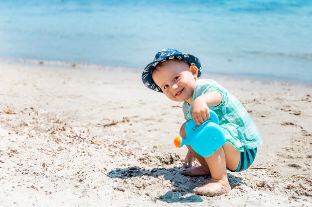 Kleiner glücklicher Kleinkindjunge spielt mit einer Spielzeuggießkanne mit Sand und Wasser am Strand.