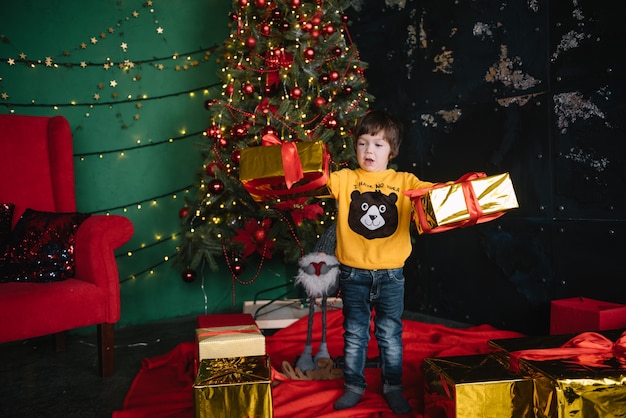 Kleiner glücklicher niedlicher Junge nahe dem Weihnachtsbaum und hält eine Geschenkbox. Kind. Weihnachtsstimmung. Ferien. Weihnachten. Neujahr.