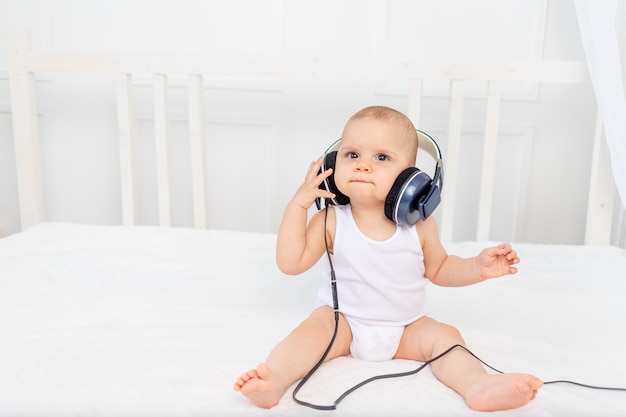 Kleiner Junge 8 Monate alt, der auf dem Bett im Kinderzimmer mit Kopfhörern sitzt und Musik hört