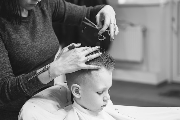 Kleiner Junge beim Friseur. Kind hat Angst vor Haarschnitten. Die Hände des Friseurs machen dem kleinen Jungen die Frisur, ganz nah. Modischer Haarschnitt für Jungen.