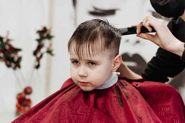 Kleiner Junge bekommt sein Haar mit einer Friseurschere geschnitten Nahaufnahme von Frauenhänden, die das Haar eines Jungen im Friseursalon pflegen Portrait eines männlichen Kindes im Friseursalon, um seine Haare zu schneiden