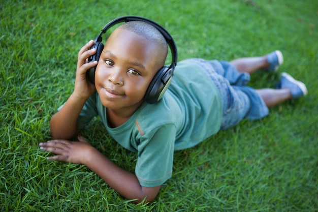 Kleiner Junge, der auf dem Gras hört Musik hört an der Kamera liegt