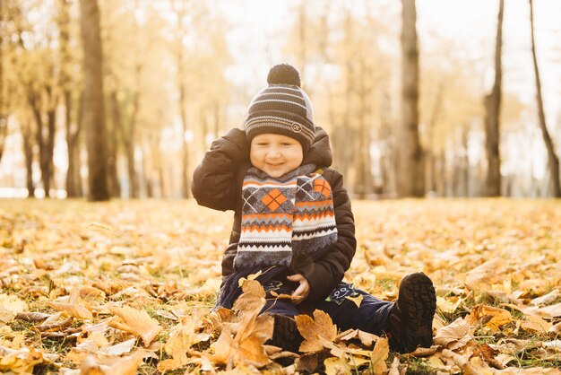 Kleiner Junge, der auf den gelben Blättern im Herbstpark sitzt