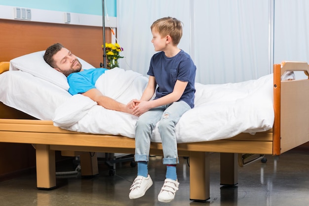 Kleiner Junge, der auf einem Krankenhausbett sitzt und den kranken Vater, Vater und Sohn im Krankenhaus ansieht