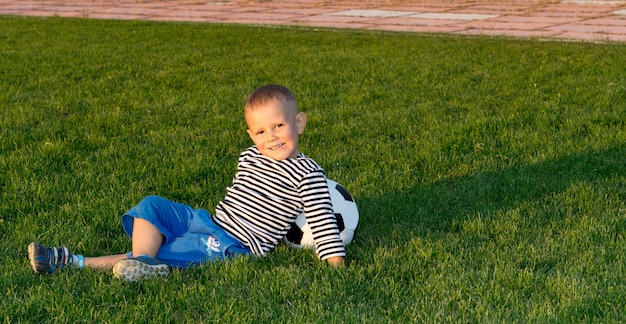 Kleiner Junge, der auf grünem Gras im Abendsonnenlicht liegt, das sich gegen einen Fußball zurücklehnt