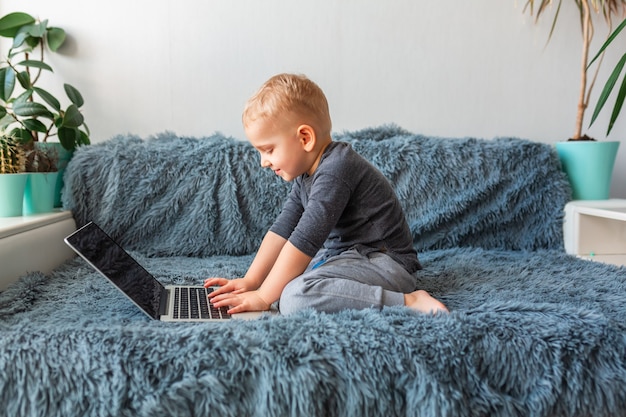 Kleiner Junge, der auf Laptop auf Sofa zu Hause spielt. E-Learning, Fernstudium, Fernkommunikationskonzept