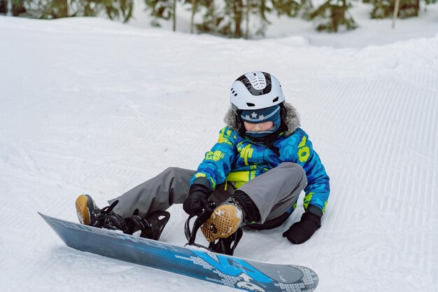 Kleiner Junge, der auf Schnee sitzt und seine Füße in Snowboardbindungen steckt und die Riemen anpasst