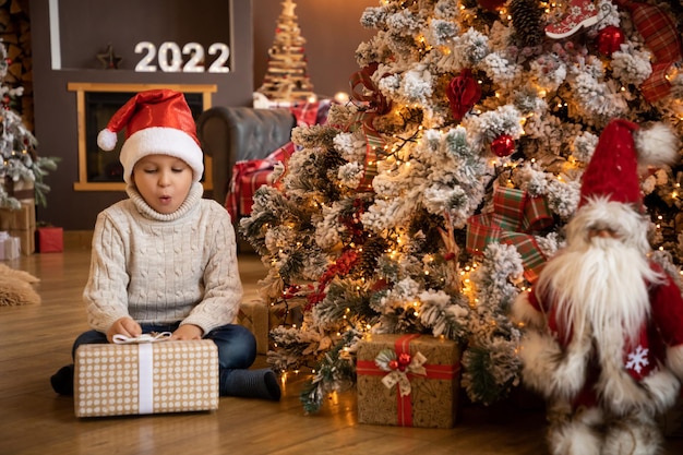 Kleiner Junge, der ein Geschenk in der Hand hält und zu Hause neben dem Weihnachtsbaum sitzt Frohes neues Jahr