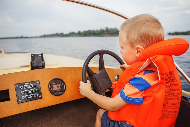 Kleiner Junge, der ein Motorboot auf Fluss fährt, der das Lenkrad fest hält