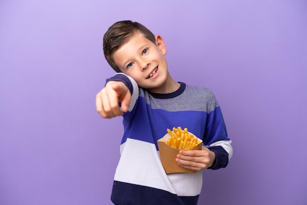 Kleiner Junge, der gebratene Chips isoliert auf violettem Hintergrund hält und mit glücklichem Gesichtsausdruck nach vorne zeigt