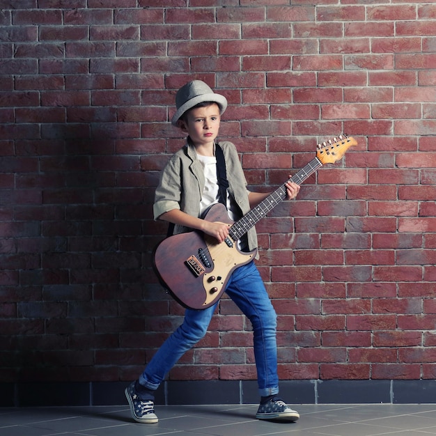 Kleiner Junge, der Gitarre auf einem Backsteinmauerhintergrund spielt