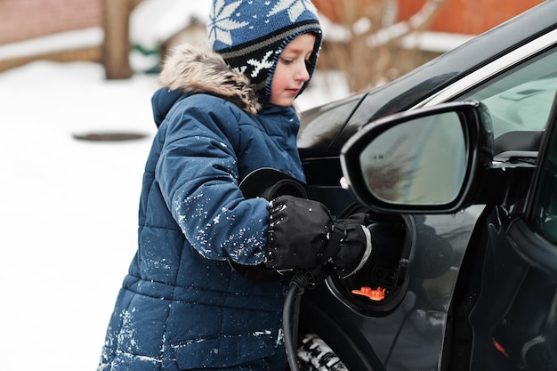 Kleiner Junge, der im Winter Elektroauto im Hof des Hauses auflädt.