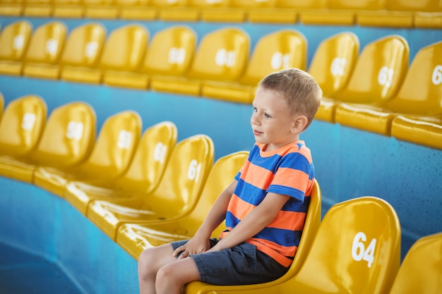 Kleiner Junge, der in einem leeren Stadion zwischen gelben Plastiksitzen sitzt