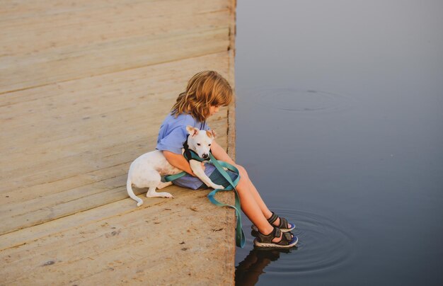 Kleiner Junge, der mit seinem Hund auf dem See sitzt, umarmt das träumende Welpenkind