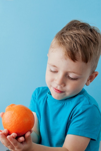 kleiner Junge, der Orange in den Händen an blauer Wand hält, Ernährung und gutes Gesundheitskonzept ausübt