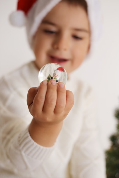 Kleiner Junge, der Weihnachtsschneeball auf dem weißen Hintergrund hält. Glückliches Kind, Weihnachtsfeier, Weihnachtszeit.