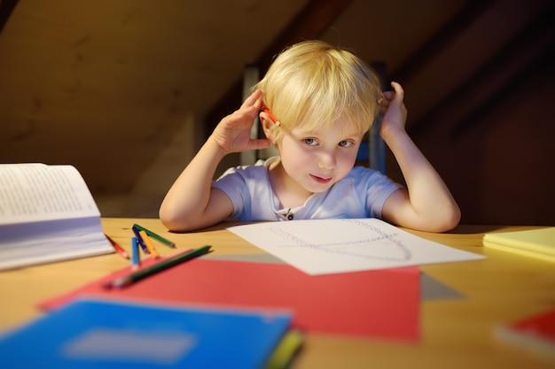 Kleiner Junge, der zu Hause Hausarbeit tut, malt und Abend schreibt. Vorschulkind lernen Lektionen - zeichnen und Farbbild. Kindertraining zum Schreiben und Lesen.