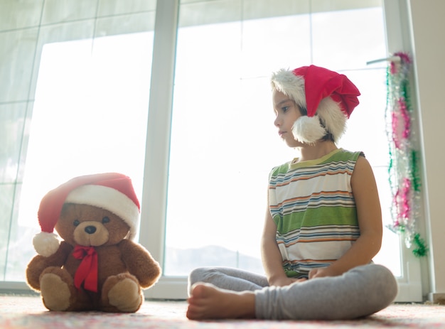 Kleiner Junge des Weihnachtsbildes in Sankt-Hut mit dem netten Teddybären, der vor Fenster sitzt