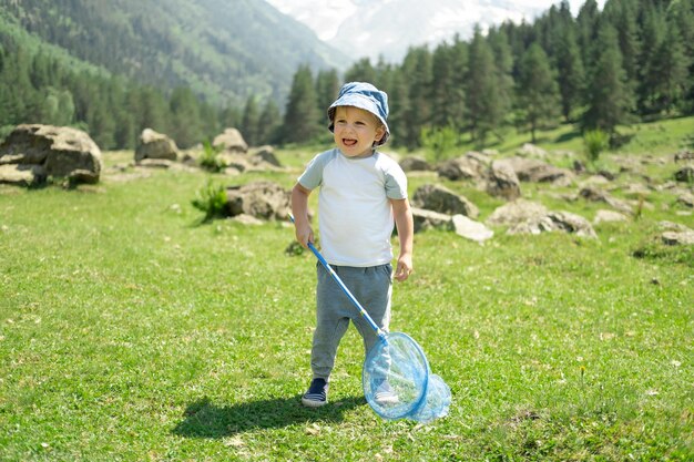 Kleiner Junge geht mit Schmetterlingsnetz spazieren und fängt Schmetterlinge auf grünen Hügeln an sonnigen Sommertagen