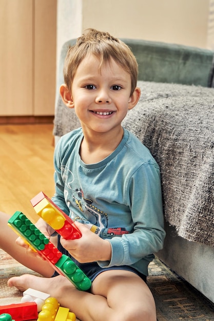 Kleiner Junge hält bunte Konstrukteursdetails aus Kunststoff in der Hand und sitzt auf dem Boden neben dem Sofa im Wohnzimmer