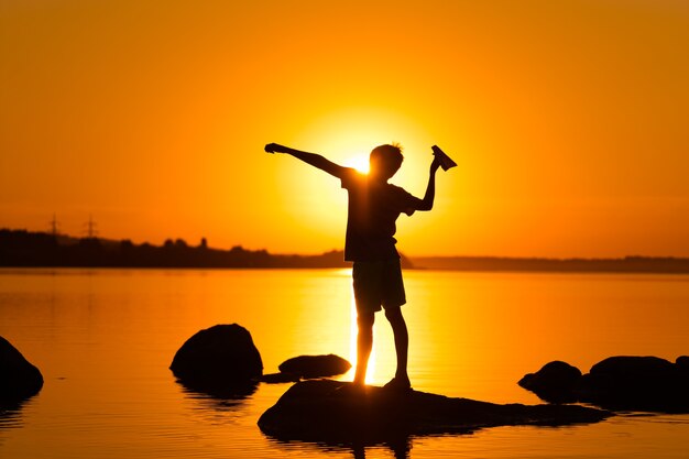 Kleiner Junge hält einen Papierflieger in der Hand am Fluss. Schöner orangefarbener Sonnenuntergang im Sommer. Silhouette eines Jungen mit Origami-Flugzeug, das in der Nähe des Sees steht.