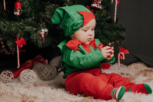 Kleiner Junge im Elfenkostüm sitzt unter einem Weihnachtsbaum mit Bällen