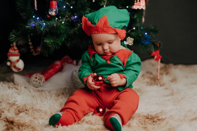 Kleiner Junge im Elfenkostüm sitzt unter einem Weihnachtsbaum mit Bällen