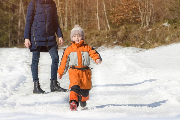 Kleiner Junge im Overall läuft von seiner Mutter weg. Verschneiter Wintertag im Nadelwald.