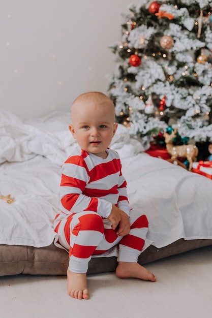 Kleiner Junge im rot-weißen Schlafanzug sitzt auf der Bettkante neben dem Weihnachtsbaum