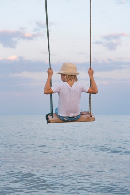 Kleiner Junge im Strohhut, der auf Schaukel über dem Wasser reitet. Sommerferien.