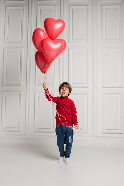 Kleiner Junge in den Jeans und im Pullover, die Spaß mit roten Luftballons auf weißem Hintergrund mit Platz für Text haben