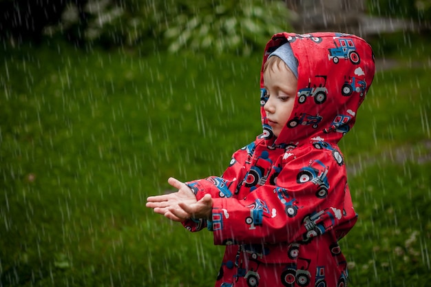 Kleiner Junge in einer wasserdichten Jacke in Traktoren fängt den Regen. Kind, das Spaß draußen in der Sommerdusche hat