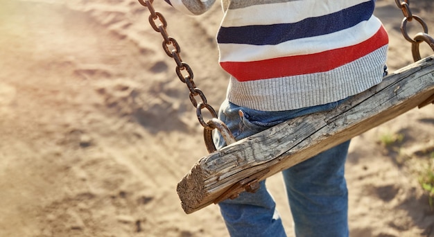 Kleiner Junge in gestreiftem Pullover und Jeans spielt bei Sonnenuntergang alte Holzschaukeln auf Sandspielplatz